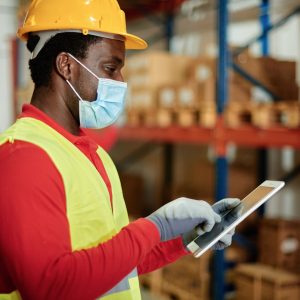 Caucasian adult warehouse worker checks delivery order on a tablet while wearing a safety mask